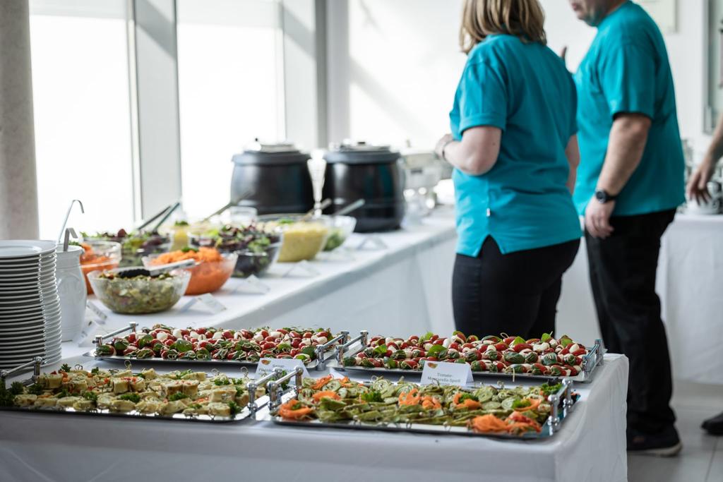 Buffet mit verschiedenen Speisen, zwei Personen in blauen Shirts stehen daneben.